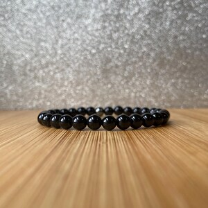 K&ouml;nnte beinhalten: Ein schwarzes Perlenarmband mit einem silbernen Verschluss. Das Armband besteht aus runden, glatten Perlen und liegt auf einer Holzoberfl&auml;che.