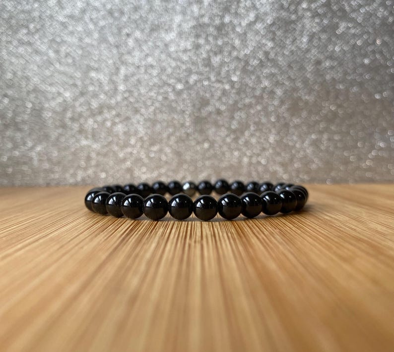 K&ouml;nnte beinhalten: Ein schwarzes Perlenarmband mit einem silbernen Verschluss. Das Armband besteht aus runden, glatten Perlen und liegt auf einer Holzoberfl&auml;che.
