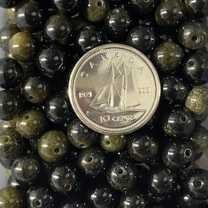 K&ouml;nnte beinhalten: Nahaufnahme eines Haufens schwarzer und gr&uuml;ner Edelsteinperlen. Die Perlen sind rund und haben eine glatte, polierte Oberfl&auml;che. Eine kanadische 10-Cent-M&uuml;nze liegt zur Gr&ouml;&szlig;enbestimmung auf den Perlen.