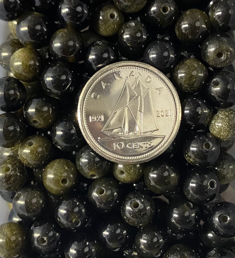 K&ouml;nnte beinhalten: Nahaufnahme eines Haufens schwarzer und gr&uuml;ner Edelsteinperlen. Die Perlen sind rund und haben eine glatte, polierte Oberfl&auml;che. Eine kanadische 10-Cent-M&uuml;nze liegt zur Gr&ouml;&szlig;enbestimmung auf den Perlen.