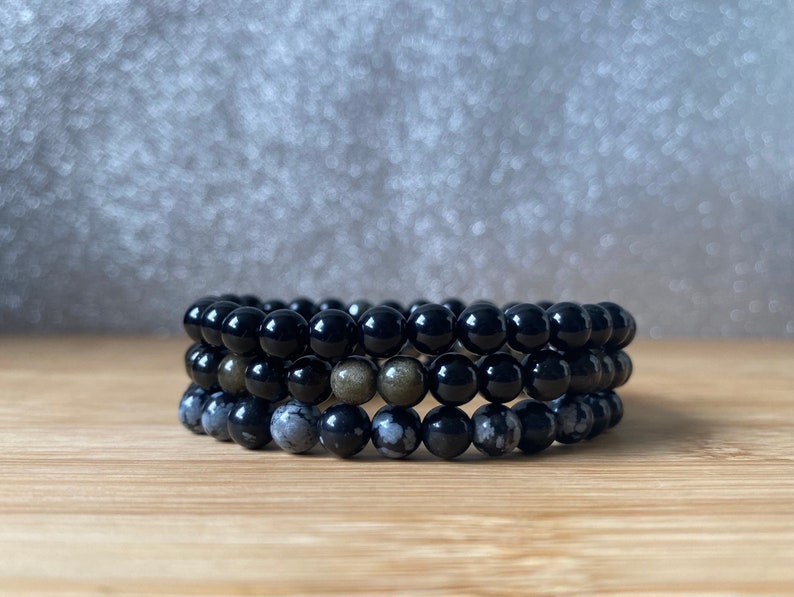 K&ouml;nnte beinhalten: Drei &uuml;bereinanderliegende Armb&auml;nder aus schwarzen, grauen und goldenen Perlen. Die Armb&auml;nder sind aus nat&uuml;rlichen Steinen.