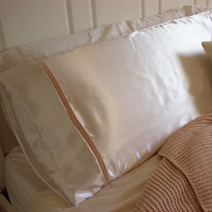 May include: A white satin pillowcase with a pink satin trim. The pillowcase is lying on a bed with a pink knitted blanket.