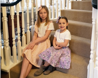 May include: Two girls wearing white t-shirts and floral skirts sit on a carpeted staircase. The girl on the left is wearing a pink and white polka dot skirt and a brown headband. The girl on the right is wearing a purple and white floral skirt and a pink bow headband.