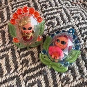May include: Two decorative pins featuring small doll figures encased in clear plastic domes. One pin has an orange floral border with gold hair, while the other has a blue floral border with pink hair. Both feature green leaf accents.