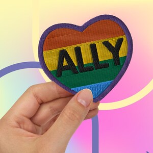 May include: A rainbow heart-shaped patch with the word "ALLY" embroidered in black letters. The patch is held in a hand against a colorful background.