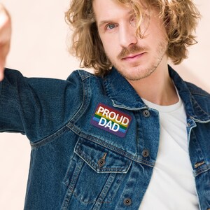 Op de afbeelding: Een donkerblauwe spijkerjas met een rechthoekige patch. De patch zegt "PROUD DAD" in witte en gouden letters, met een regenboogstreepontwerp. De jas heeft twee borstzakken en knopen.