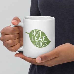 Pode incluir: Caneca de cerâmica branca com um design de folha verde e o texto "HOT LEAF JUICE" impresso nela.