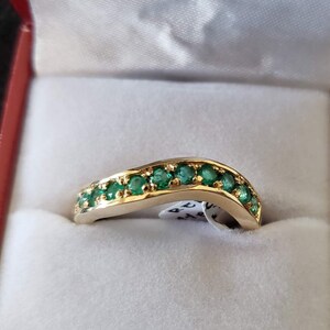 May include: A gold band ring with a row of emerald green gemstones set in a curved design. The ring is displayed in a red velvet ring box.