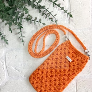 Op de afbeelding: Oranje gebreide telefoonhoes met een bijpassend bandje en een zilveren sluiting. De hoes heeft een "Handmade"-label.