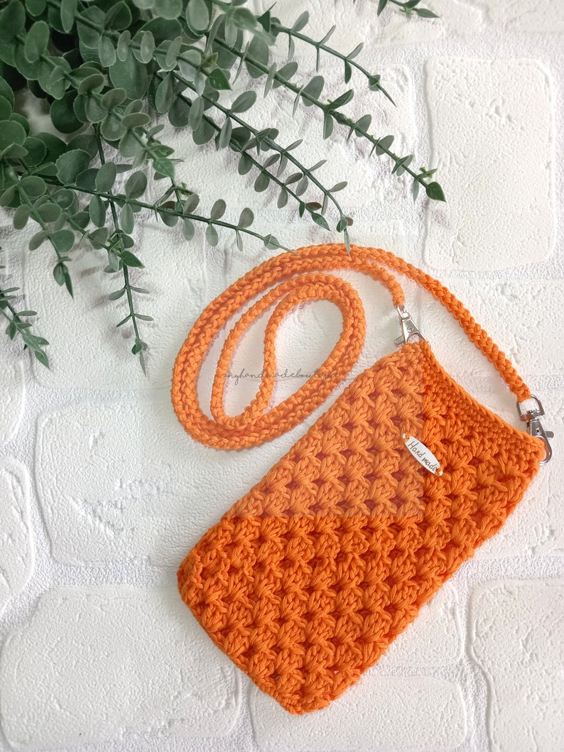 Op de afbeelding: Oranje gebreide telefoonhoes met een bijpassend bandje en een zilveren sluiting. De hoes heeft een "Handmade"-label.
