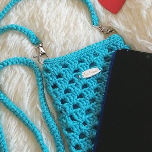 Op de afbeelding: Een turquoise gehaakte telefoonhoes met een bandje en een "Hand made" label. De hoes ligt op een wit, pluche oppervlak met een rode hartvormige uitsnijding.