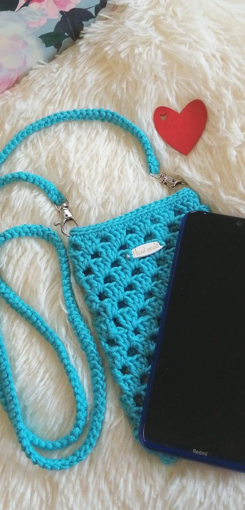 Op de afbeelding: Een turquoise gehaakte telefoonhoes met een bandje en een "Hand made" label. De hoes ligt op een wit, pluche oppervlak met een rode hartvormige uitsnijding.