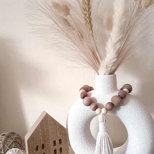 May include: A white ceramic vase with a brown wooden bead garland and a white tassel. The vase is filled with dried pampas grass and other dried flowers.