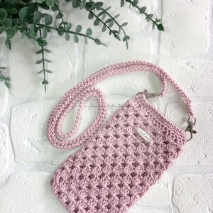 Op de afbeelding: Een roze gehaakte telefoonhoes met een lange band. De hoes heeft een klein label met de tekst "Handmade".