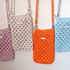 Op de afbeelding: Vier gehaakte mini-tasjes met verstelbare banden die aan een witte muur hangen. De tasjes zijn roze, cr&egrave;me, oranje en lichtblauw. Elk tasje heeft een klein wit label met de tekst "Hand Made".