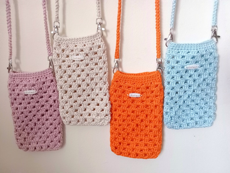 Op de afbeelding: Vier gehaakte mini-tasjes met verstelbare banden die aan een witte muur hangen. De tasjes zijn roze, cr&egrave;me, oranje en lichtblauw. Elk tasje heeft een klein wit label met de tekst "Hand Made".