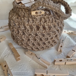 May include: A brown jute rope basket with a wooden button closure. The basket is filled with wooden clothes pegs. The basket is sitting on a white surface.