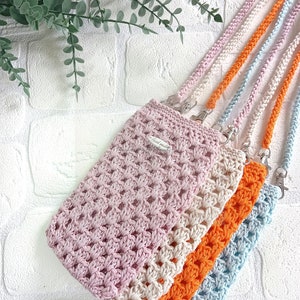 Op de afbeelding: Vier gehaakte telefoonhoesjes in roze, cr&egrave;me, oranje en blauw met bijpassende gehaakte bandjes. De hoesjes zijn op elkaar gestapeld en het roze hoesje heeft een "Hand made"-label.