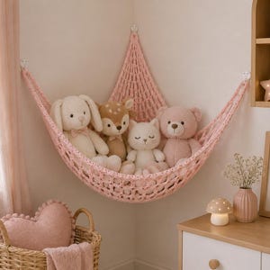 Op de afbeelding: Een roze macramé hangmat met vier knuffels: een witte konijn, een bruin hert, een witte kat en een roze beer. De hangmat hangt aan een lichtroze muur in een kinderkamer. Een hartvormig kussen en een rieten mand staan op de voorgrond.
