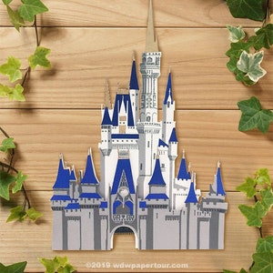 May include: A papercraft model of Cinderella's Castle, a white castle with blue accents and gold trim, against a woodgrain background. The castle has multiple towers and a pointed roof. The text "©2019 wdwpapertour.com" is at the bottom of the image.
