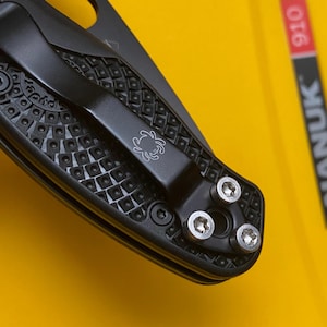 May include: Close-up of a black folding knife with a textured handle and a pocket clip featuring a spider logo. The knife has three silver screws and a decorative pattern on the handle. The background is yellow.