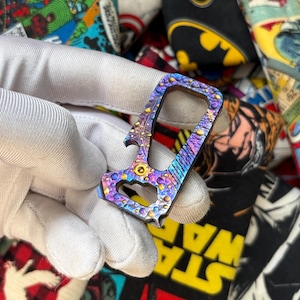 May include: A multi-tool with a colorful, iridescent finish. The tool features a bottle opener, a pry bar, and other functional elements. The design incorporates a floral pattern and a textured surface, with shades of purple, blue, and gold.