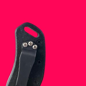 May include: Close-up of a black pocket knife handle with a textured grip. The handle features a metal pocket clip and is secured with three small screws. A lanyard hole is visible at the top of the handle.