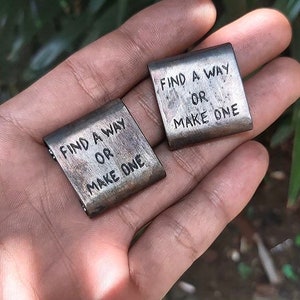 May include: Two small, rectangular metal tags with the words "Find a way or make one" engraved on them. The tags are held in a hand.