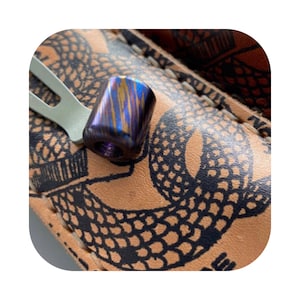 Puede incluir: Una pequeña cuenta cilíndrica hecha de titanio azul y morado con un patrón de remolino. La cuenta está sobre una superficie de cuero marrón con un diseño de serpiente negra.