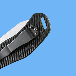 Puede incluir: Primer plano de una navaja plegable negra con un mango texturizado y una hoja plateada. El mango tiene un clip para el bolsillo y est&aacute; asegurado con tornillos negros. El cuchillo tiene un orificio para un cord&oacute;n.