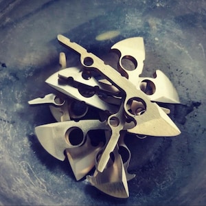 May include: A pile of six brass throwing knives with intricate cutouts and sharp points. The knives are arranged in a random pattern, with some overlapping.