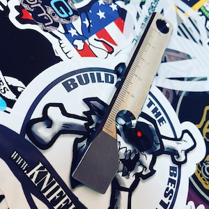 May include: A silver metal tool with a ruler on one side and a flat edge on the other. The tool is lying on top of a sticker that says "Build the Best!" with a skull and crossbones design. The sticker is on a white background.