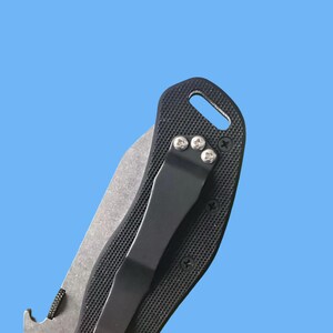 Puede incluir: Primer plano de una navaja plegable negra con mango texturizado y hoja plateada. La navaja presenta un clip de bolsillo negro y tornillos plateados. El fondo es azul claro liso.