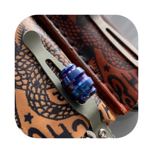 Puede incluir: Primer plano de un spinner fidget de titanio azul y morado con acabado texturizado. El spinner está unido a un clip de bolsillo plateado.