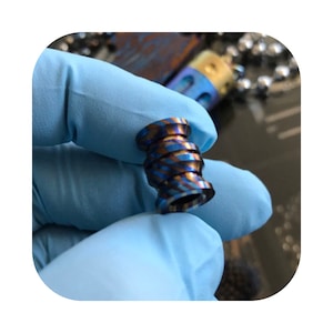 Puede incluir: Una pequeña cuenta cilíndrica con un diseño en espiral. La cuenta presenta una combinación de colores azul y dorado. La cuenta es sostenida por una mano enguantada. La cuenta es probablemente un componente de joyería o un proyecto de artesanía similar.