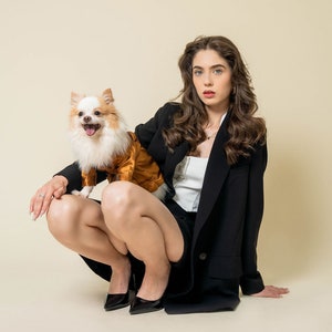 May include: A woman wearing a black blazer and a white top is sitting with a small white and brown dog wearing a gold jacket. The dog is sitting on the woman's lap.