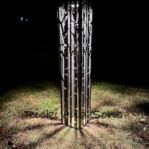 May include: A tall, rectangular, black metal outdoor lamp with a tree-like design cut into the sides. The lamp is illuminated from within, casting light onto the surrounding grass. The text "Ricki and Sows" is at the base.
