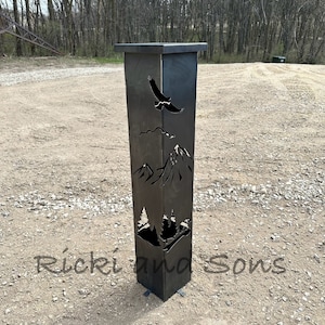 May include: A black metal fire pit with a cut-out design of mountains, trees, and an eagle. The fire pit is standing on a gravel surface. The text "Ríckiana Sons" is visible in the lower right corner of the image.