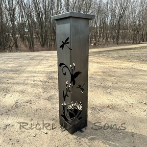 May include: A black metal garden post with a square top and cutouts of butterflies and flowers. The post is standing on a gravel driveway.