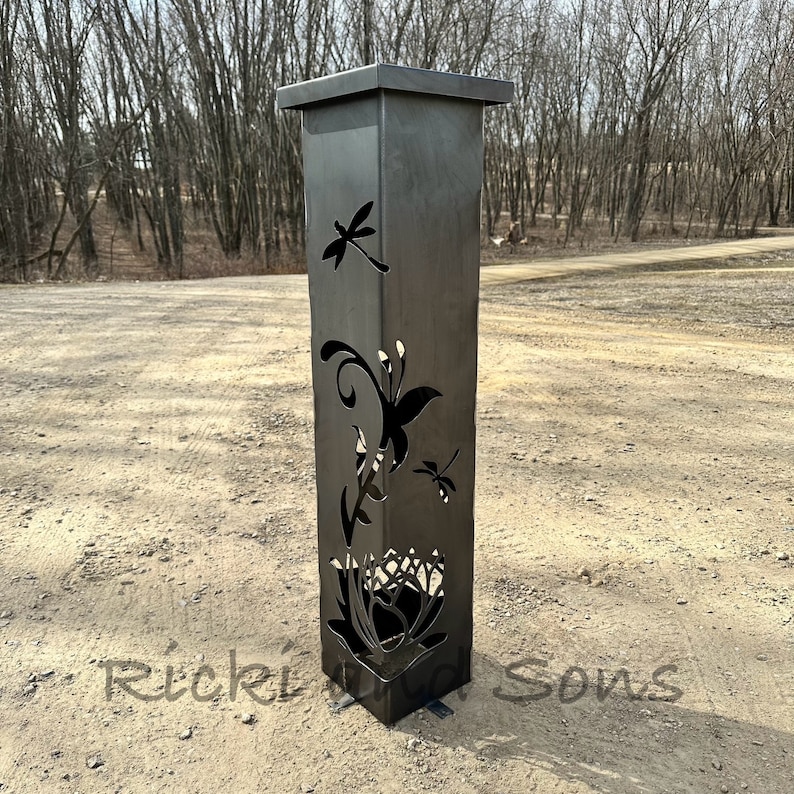 May include: A black metal garden post with a square top and cutouts of butterflies and flowers. The post is standing on a gravel driveway.