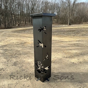 May include: A black metal garden post with a square top and a base. The post has cutouts of dragonflies and a flower design. The post is standing on a gravel surface.