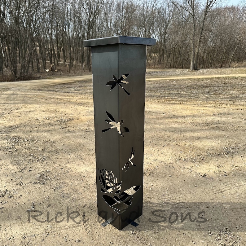 May include: A black metal garden post with a square top and a base. The post has cutouts of dragonflies and a flower design. The post is standing on a gravel surface.