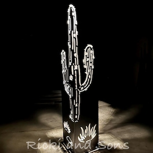 May include: A black metal cactus sculpture with white illuminated edges and details. The sculpture is mounted on a black rectangular base with a desert plant design. The base has the text "Ricki and Sons" on it.