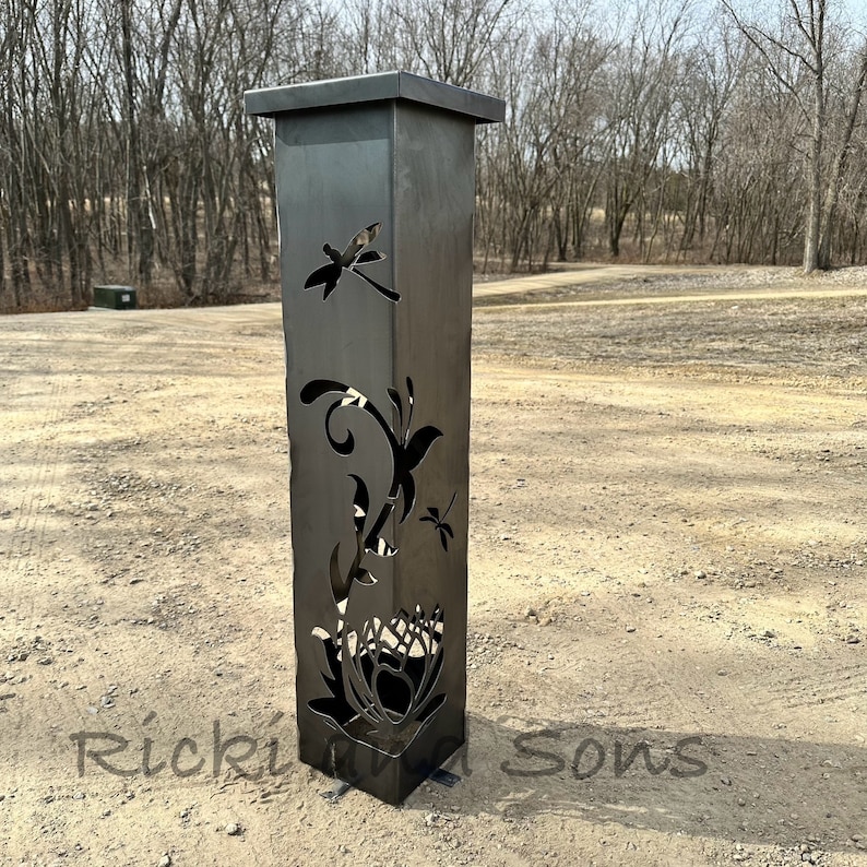 May include: A tall, black metal planter with a cutout design of flowers and dragonflies. The planter is standing on a gravel surface.