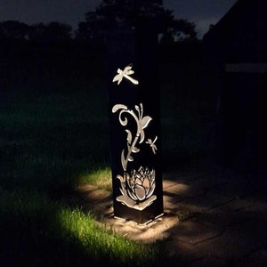 May include: A black metal garden light with a cut-out design of a dragonfly, butterfly, and flower. The light is illuminated and casting a warm glow on the surrounding grass.