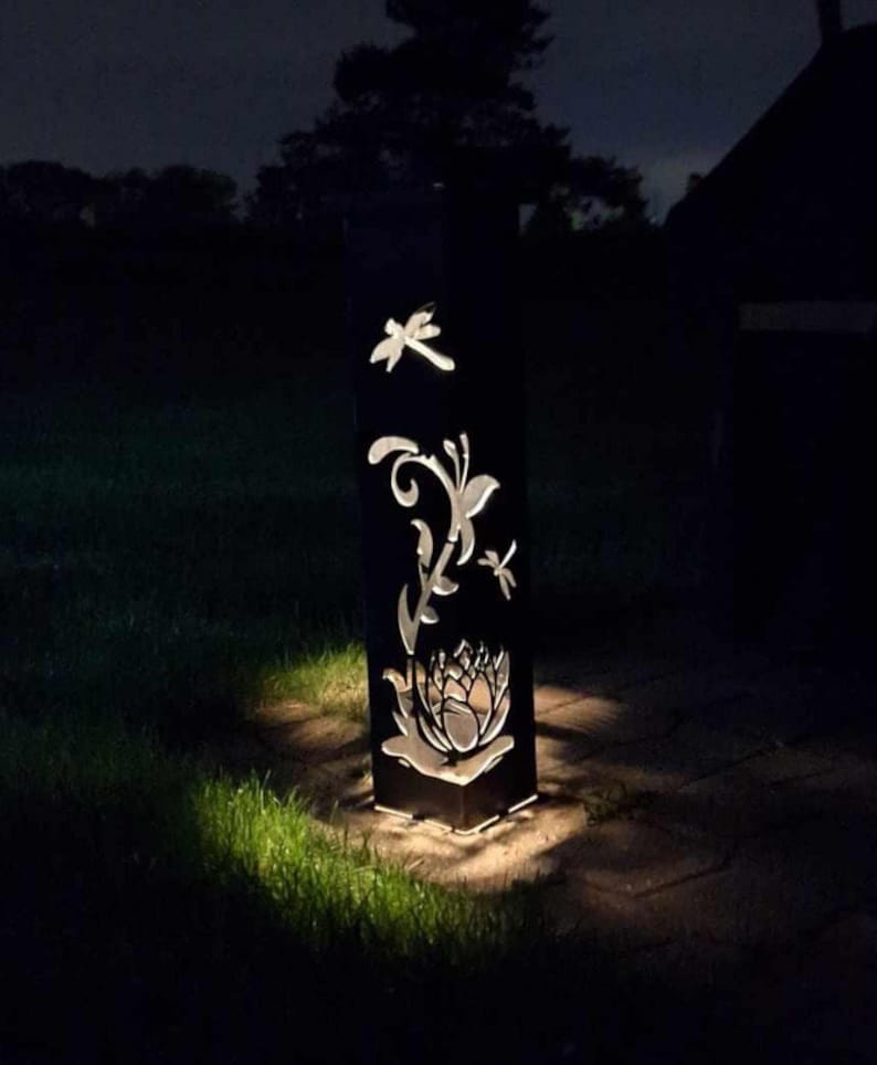 May include: A black metal garden light with a cut-out design of a dragonfly, butterfly, and flower. The light is illuminated and casting a warm glow on the surrounding grass.