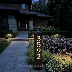 May include: A lighted address sign with the numbers 3592 illuminated in a warm glow. The sign is a dark brown rectangular post with a solar panel on top, set in a garden with lush greenery and flowers. The text "Ricki and Sons" is at the bottom.