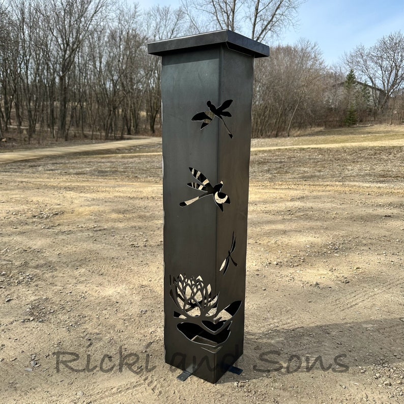 May include: A black metal garden sculpture with cutouts of dragonflies and a flower. The sculpture is standing on a gravel surface.