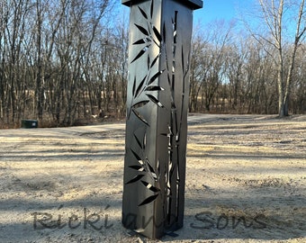 May include: A black metal outdoor lamp post with a square top and a cut-out design of bamboo leaves and stalks. The lamp post is standing on a gravel surface.