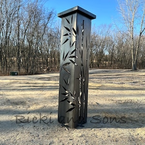 May include: A black metal outdoor lamp post with a square top and a cut-out design of bamboo leaves and stalks. The lamp post is standing on a gravel surface.